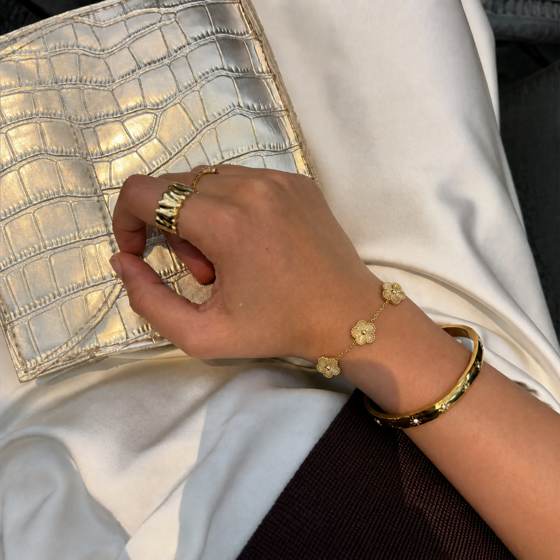 Trèfle Bracelet Gold on wrist with gold tone bangle and rings, hand resting on metallic clutch
