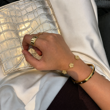 Trèfle Bracelet Gold on wrist with gold tone bangle and rings, hand resting on metallic clutch